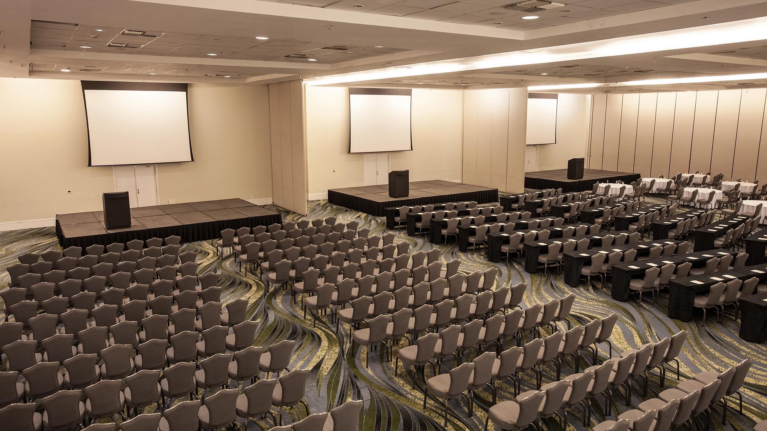 A conference room with multiple rows of chairs, a stage with a podium, and a large screen.