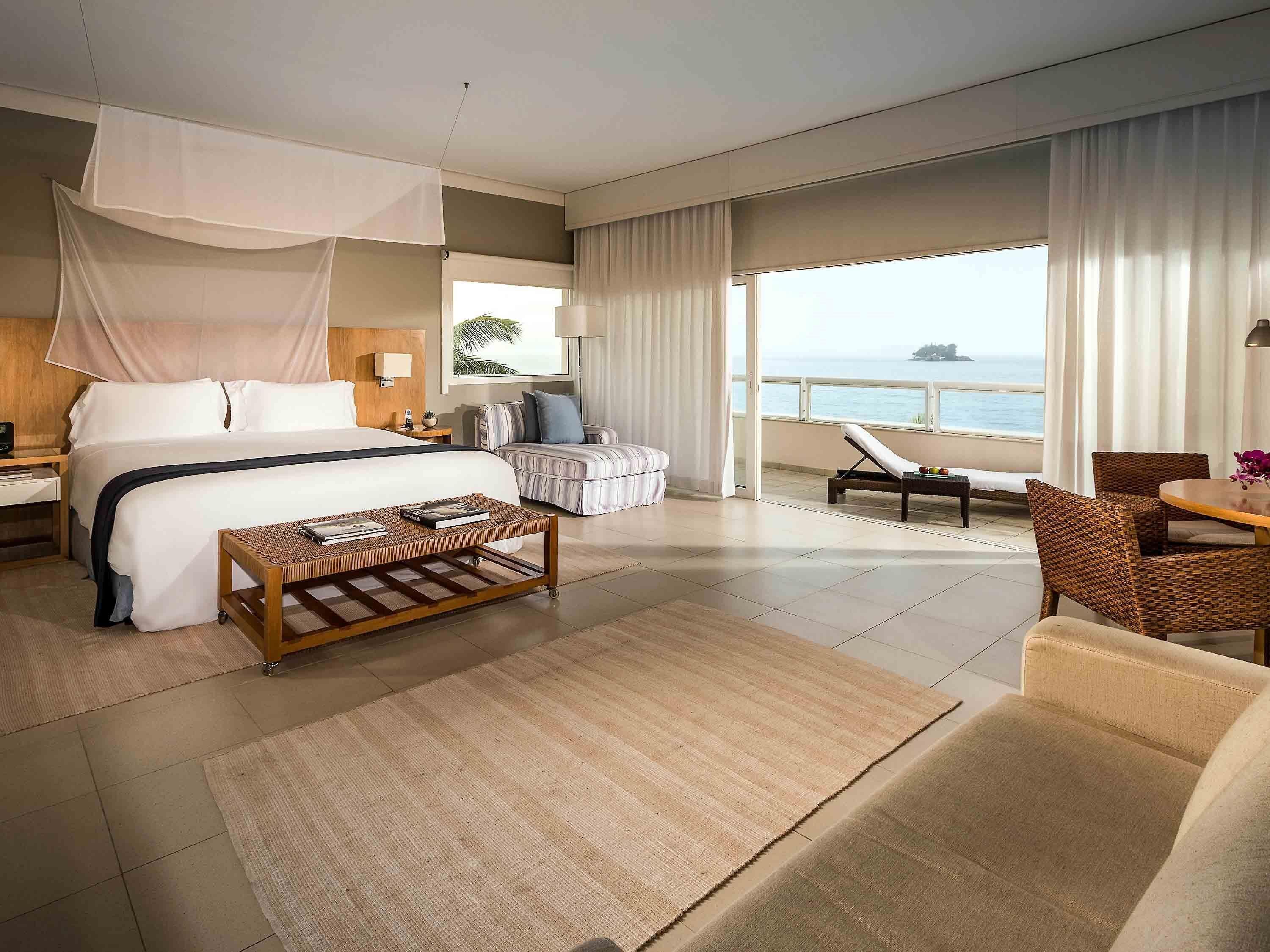 A hotel room with a bed, a coffee table, a sofa, a chair, a balcony with a view of the sea, and a window with curtains.