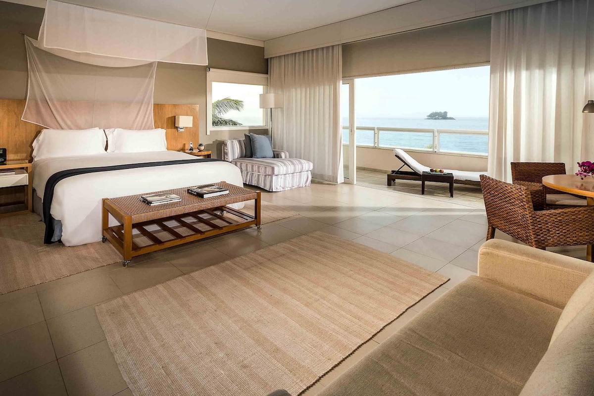 Une chambre d’hôtel comprenant un lit, une table basse, un canapé, un fauteuil, un balcon avec vue sur la mer et une fenêtre avec des rideaux.