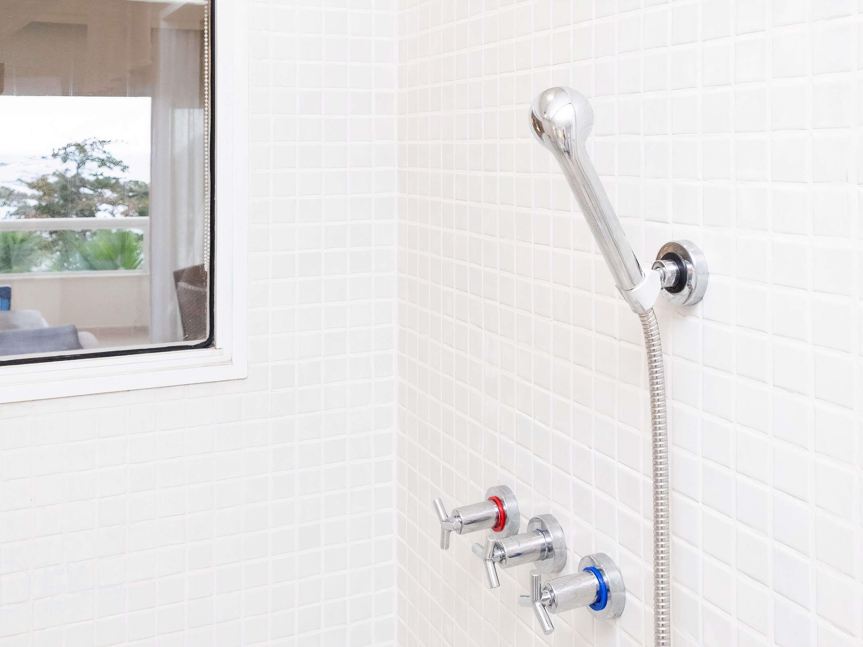 A bathroom with white tiled walls, a shower with a chrome hose, and three faucets with red, blue, and silver handles.