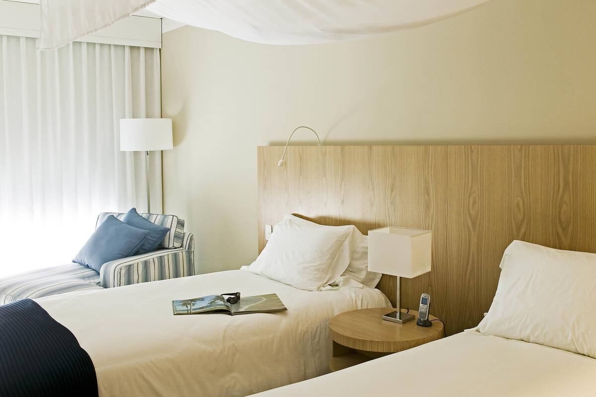 A hotel room with two beds, a bedside table with a lamp, a chair, and a window with curtains.