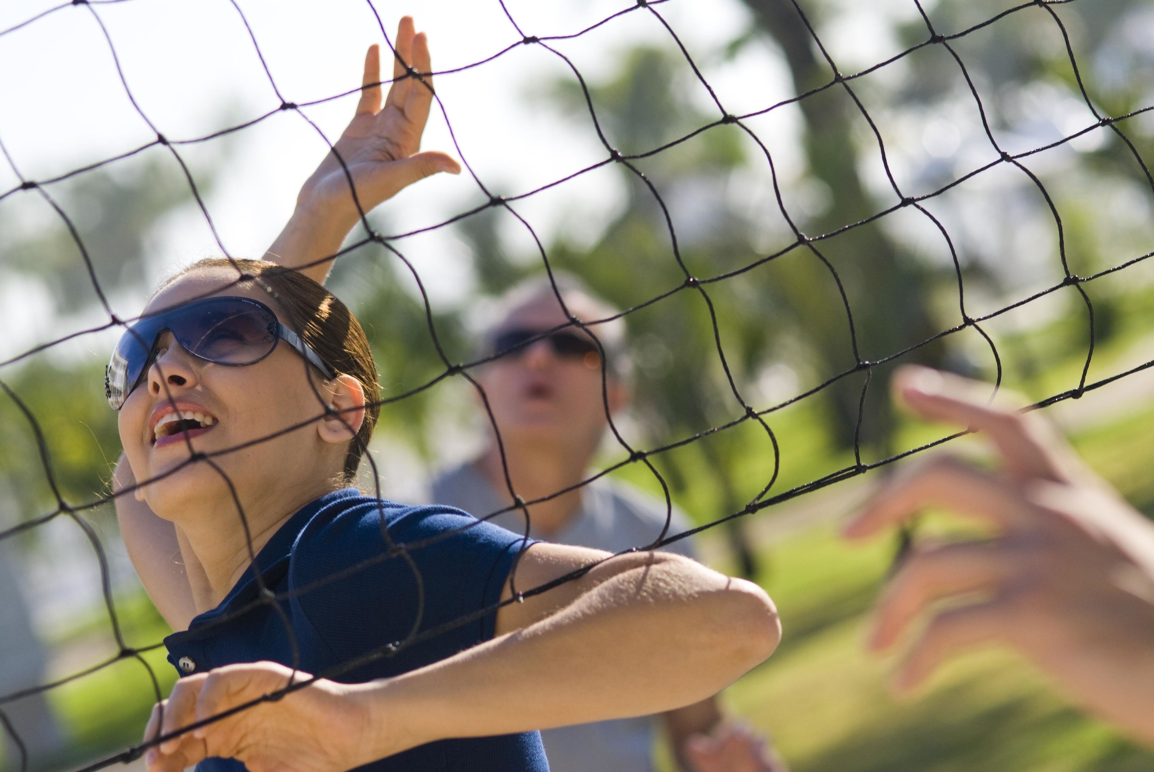 A person playing volleyball, with a net in the foreground and a blurred background of greenery.