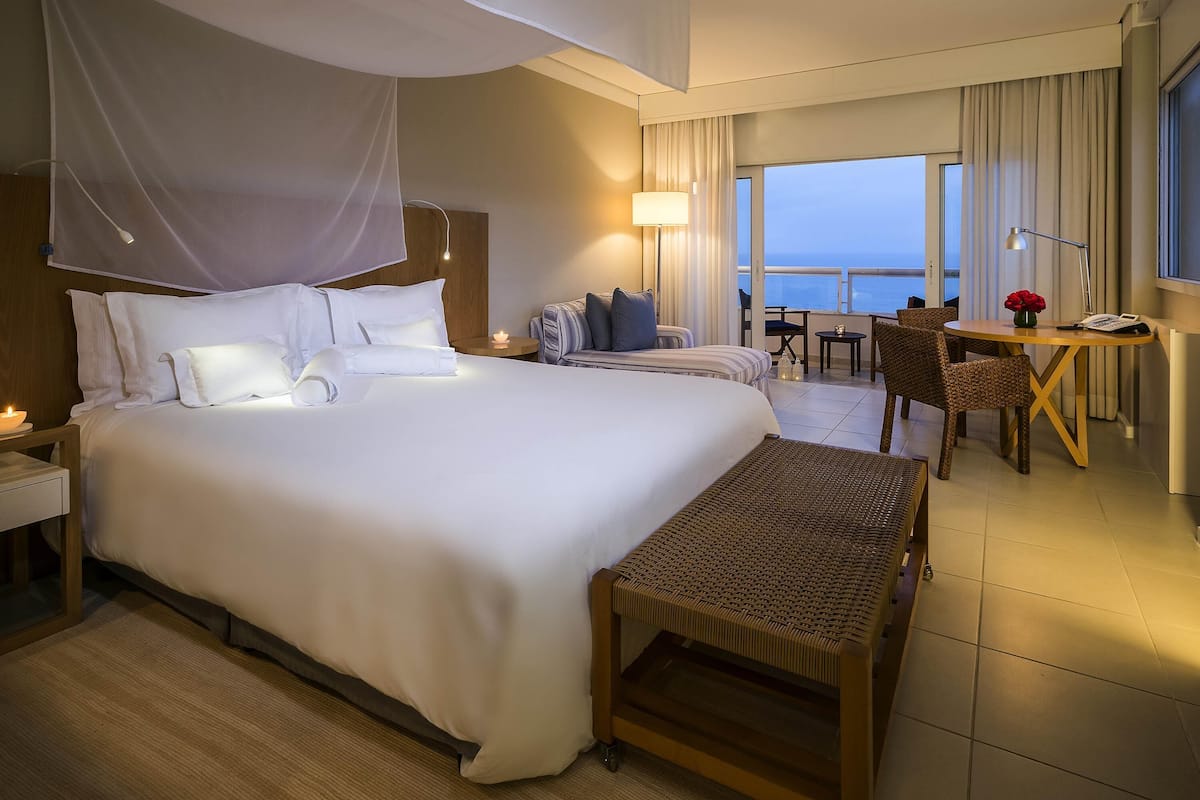 A hotel room with a large bed, a desk, a chair, a small table, and a view of the sea.