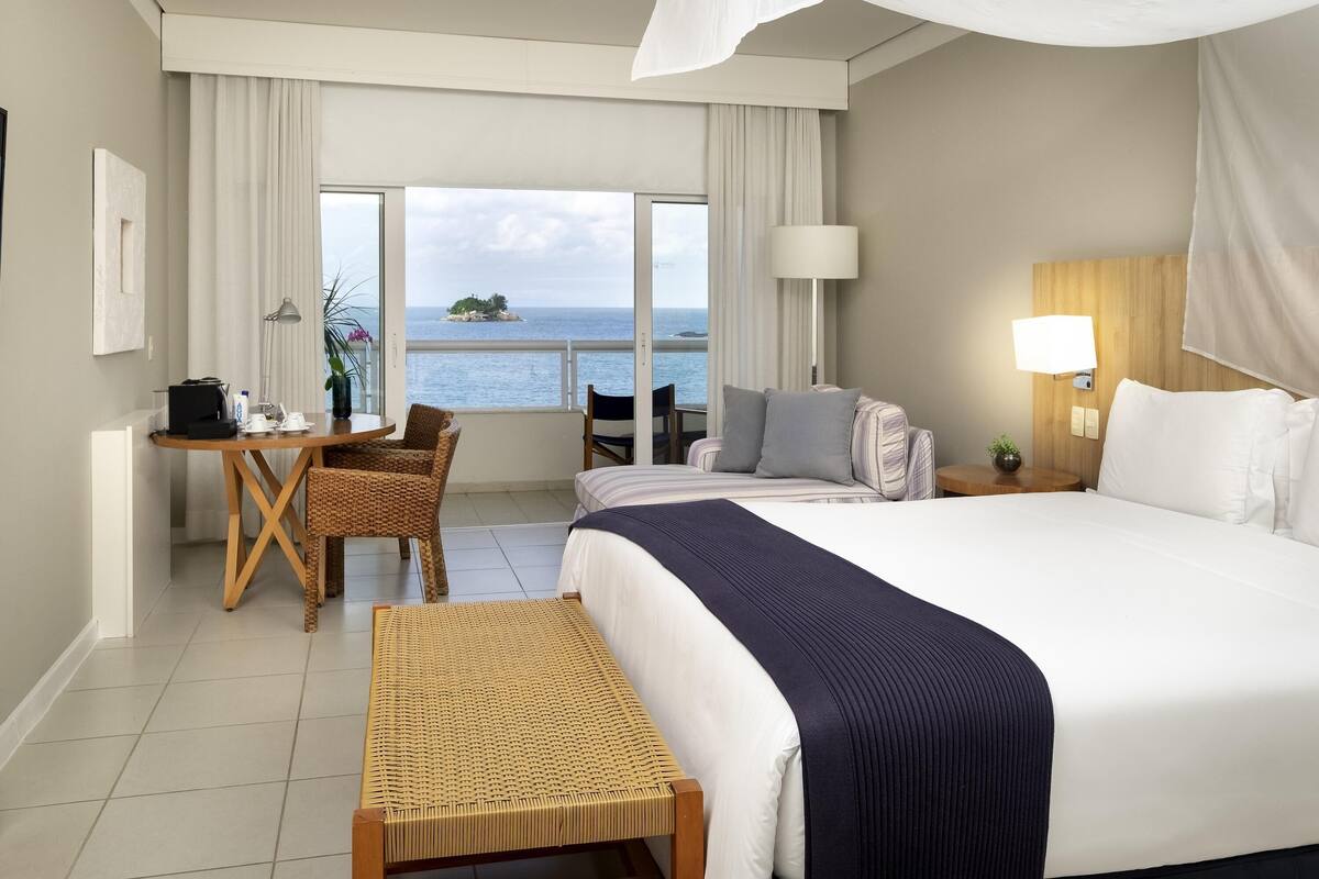 A hotel room with a bed, a desk, a chair, a small table, and a view of the sea.