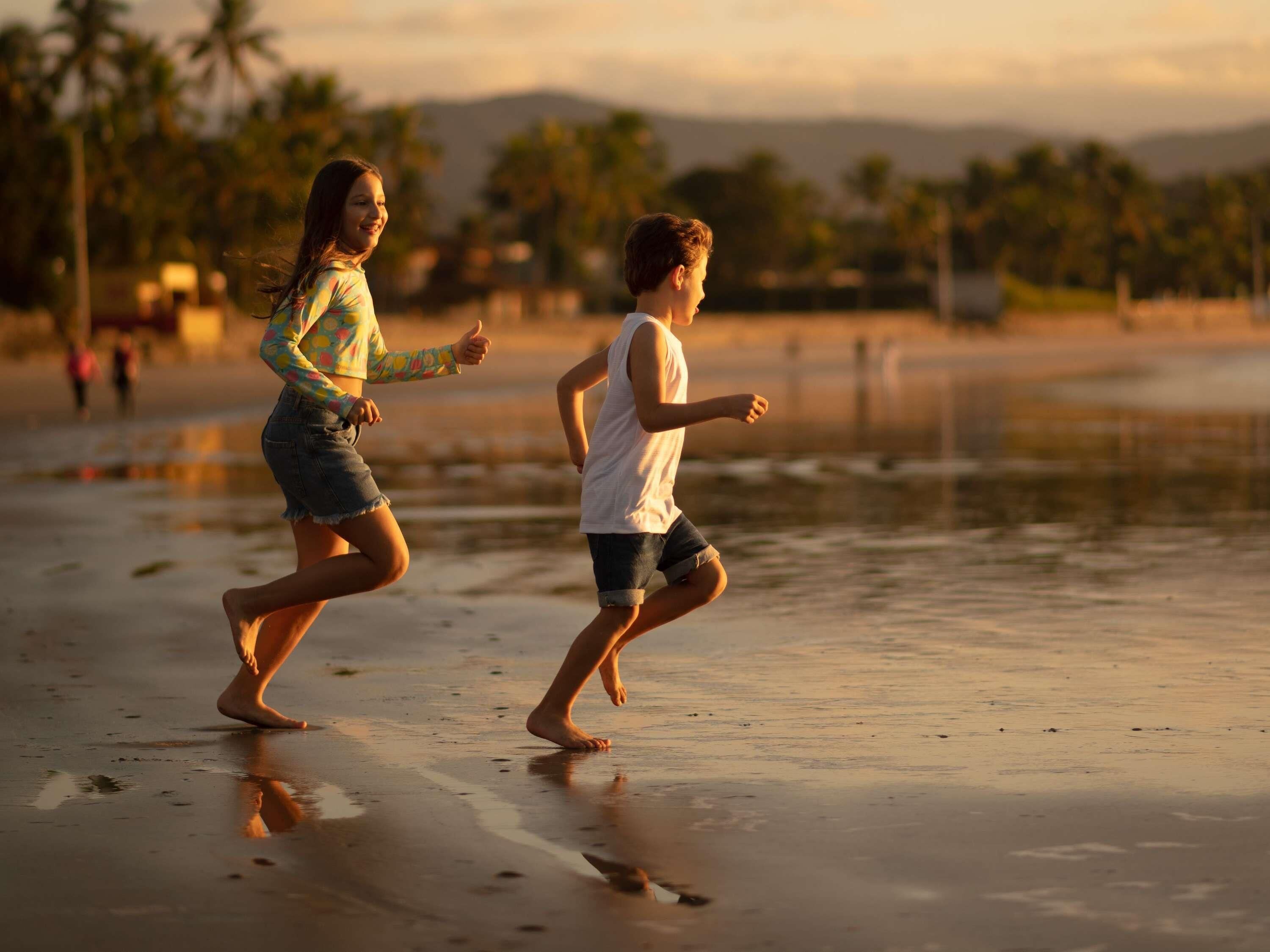 Two children running on a beach with palm trees and mountains in the background.