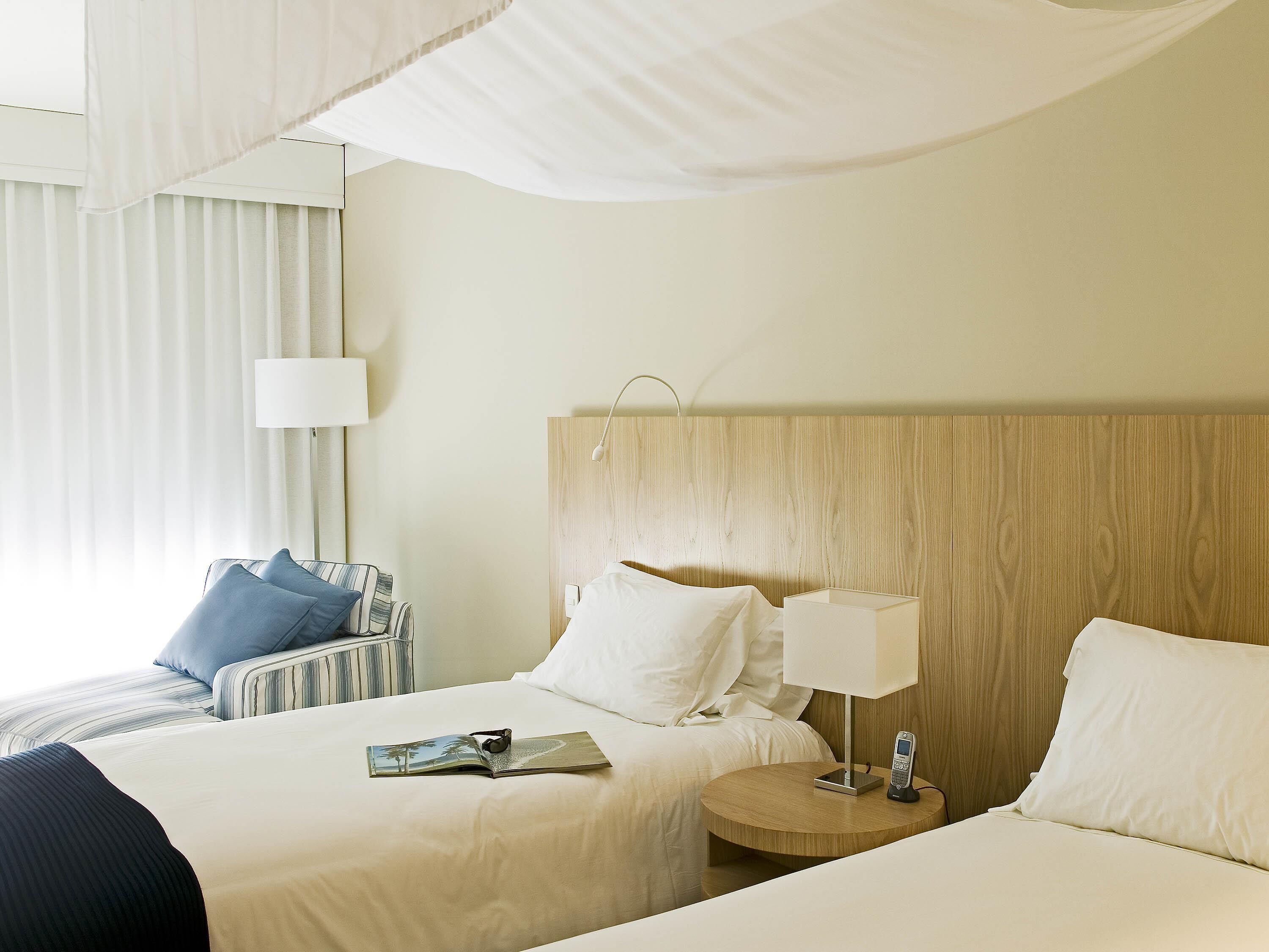 Une chambre d’hôtel avec deux lits, une table de chevet avec une lampe, une chaise et une fenêtre avec des rideaux.