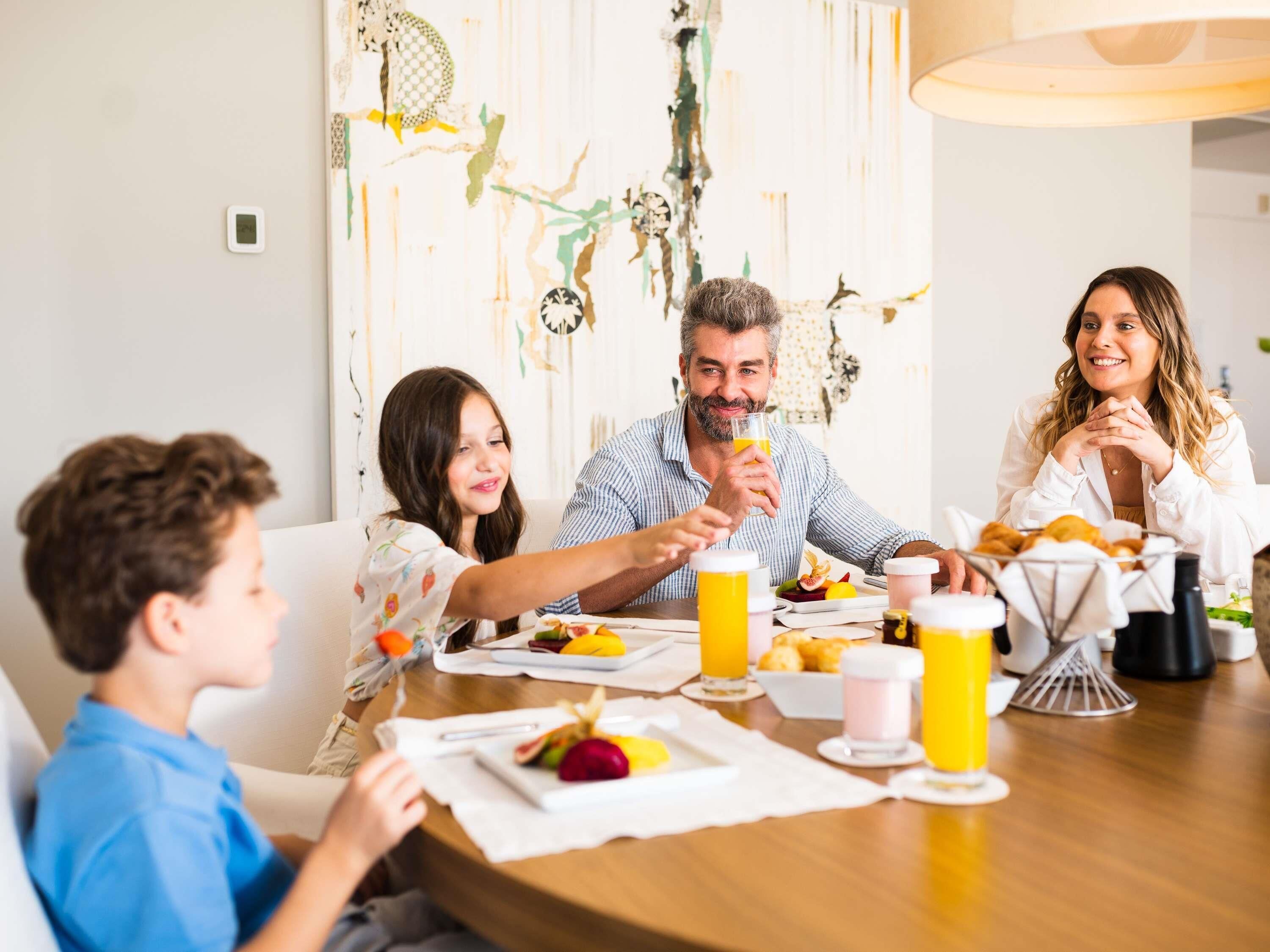 Una familia comiendo en una mesa de madera con comida y bebidas, en una habitación con arte abstracto en las paredes.