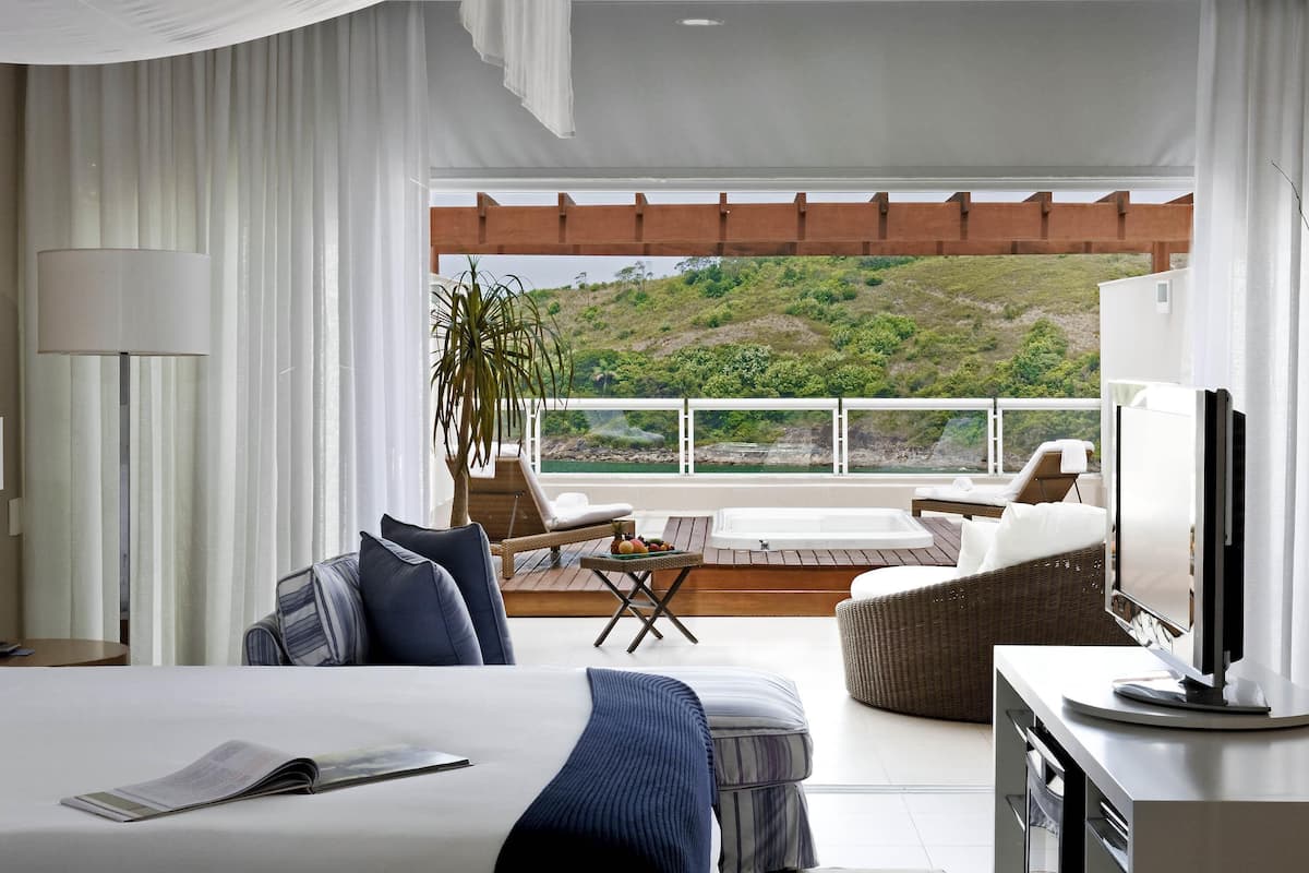A hotel room with a bed, a TV, a balcony with a view of a hillside, and a dining area with a table and chairs.