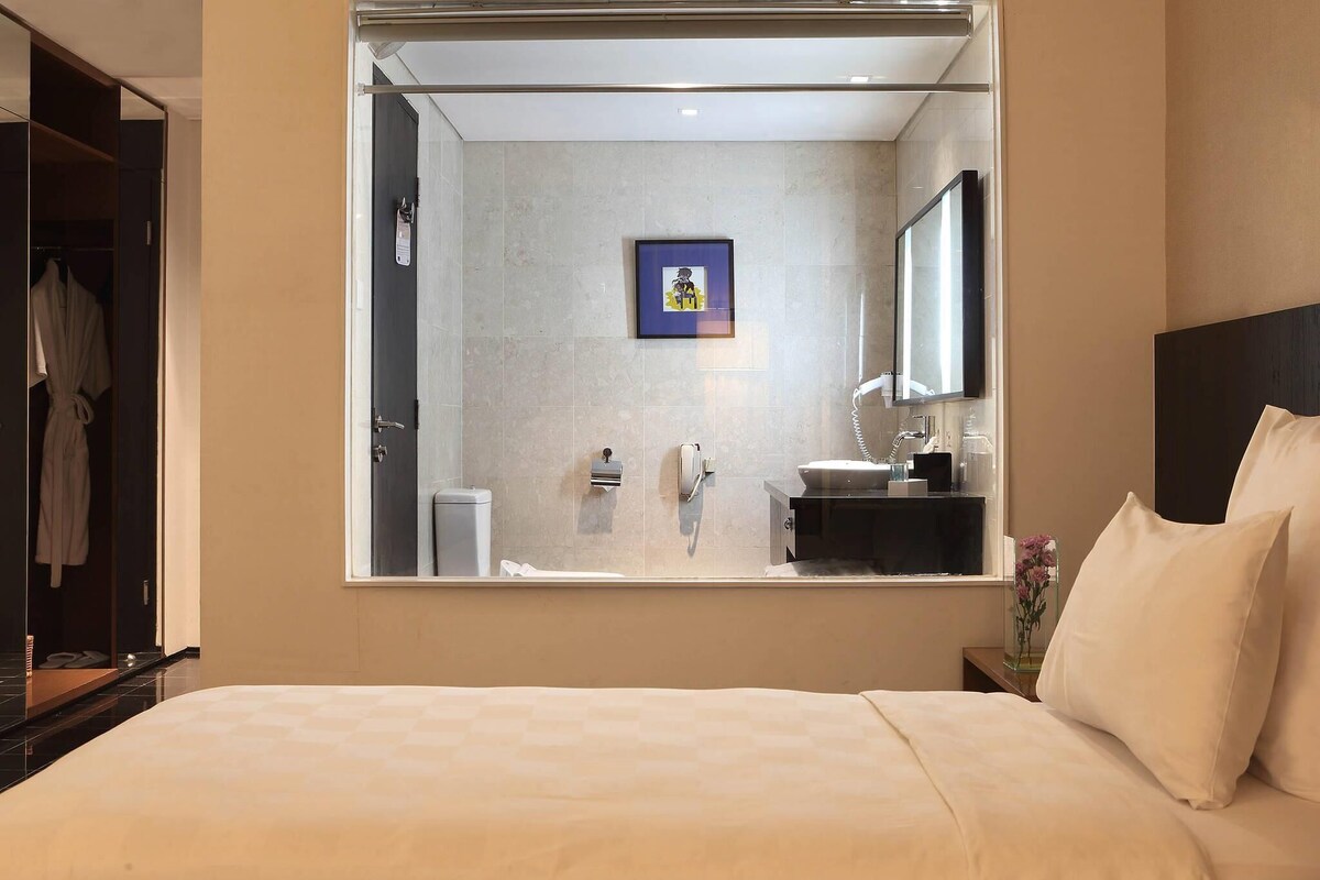 Une chambre d’hôtel avec un grand lit, une table de chevet, une commode, un miroir, une salle de bain avec un lavabo et des toilettes, et une armoire.