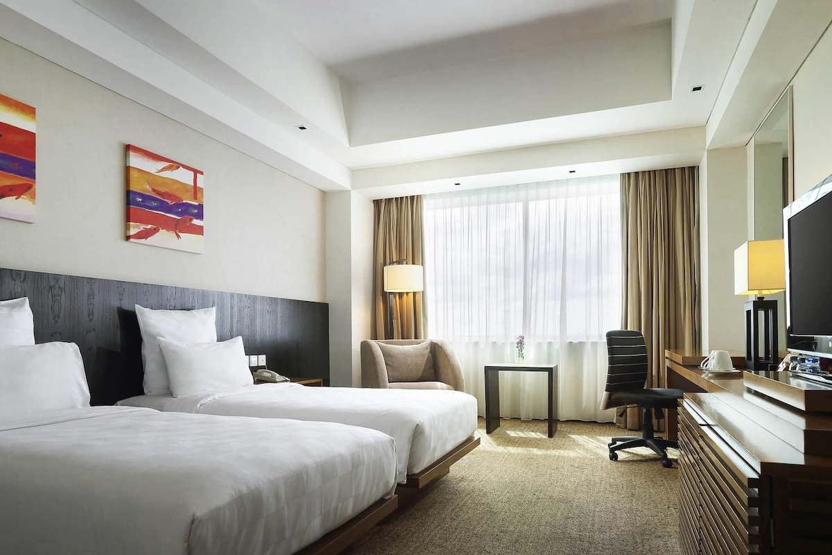 A hotel room with two beds, a desk with a chair, a TV, and a window with curtains.
