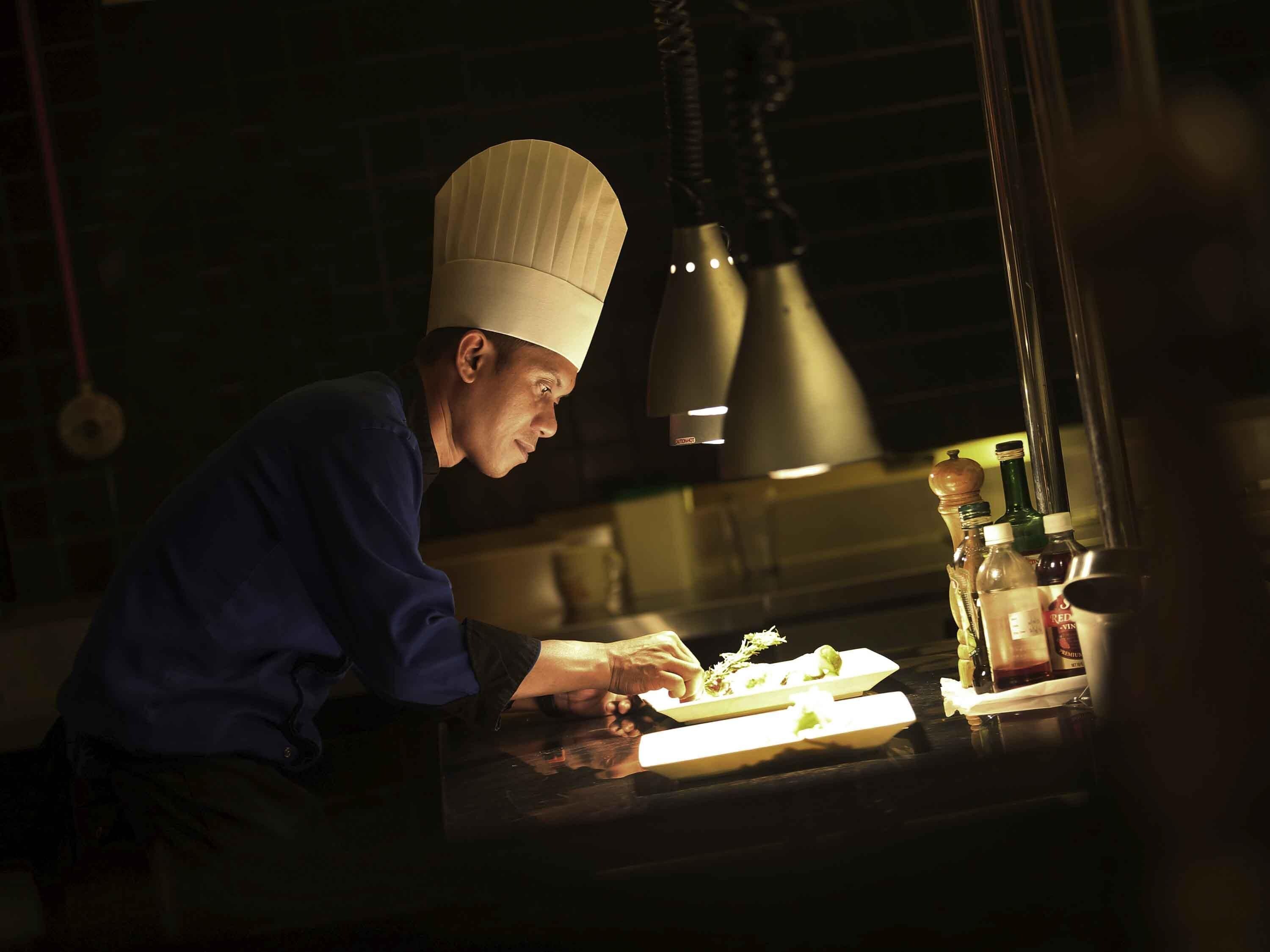 A chef in a white hat and blue uniform preparing food at a counter with a spotlight above.