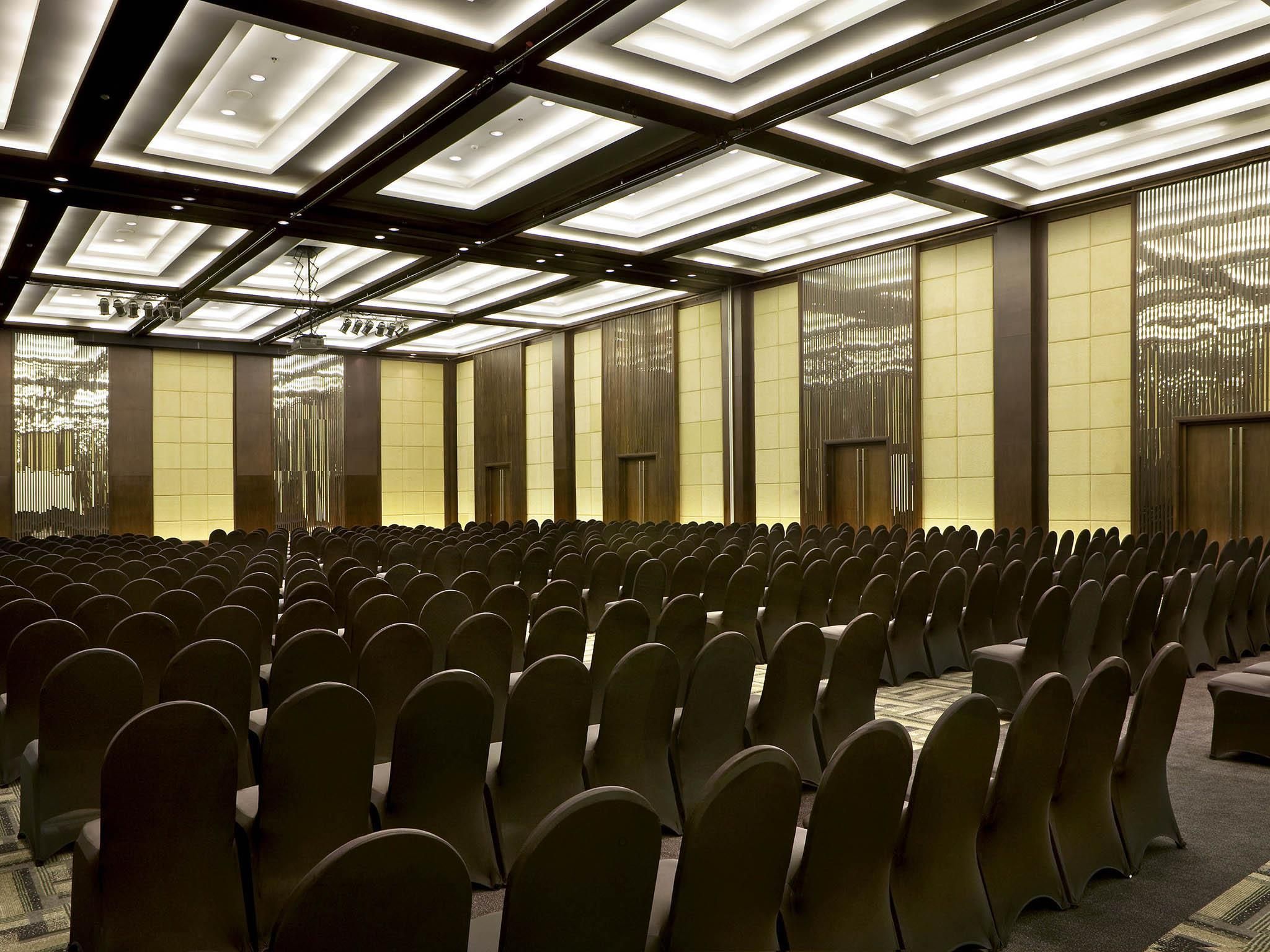 Ein Konferenzraum mit Reihen von Stühlen, einer hohen Decke mit eingelassener Beleuchtung und einem großen Fenster mit vertikalen Jalousien.