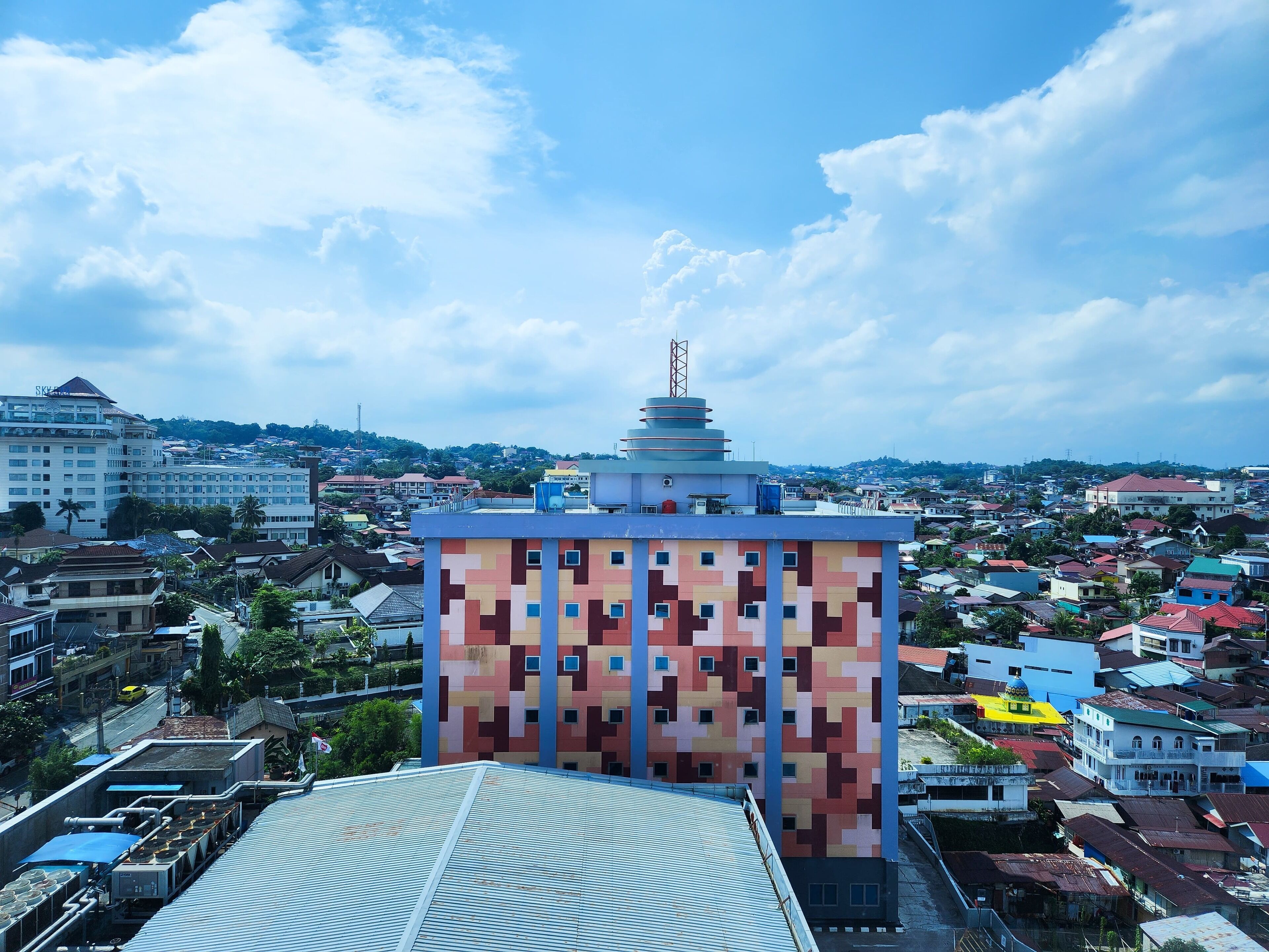 Un edificio de varios pisos con una fachada distintiva pixelada, rodeado por una zona urbana densamente poblada con varios edificios y un cielo despejado.