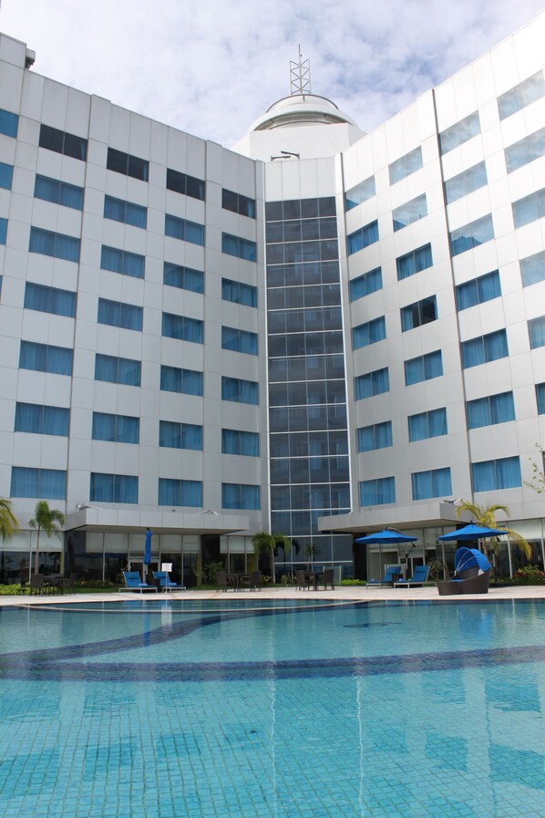 A modern hotel with a large pool area, multiple balconies, and a prominent glass facade.