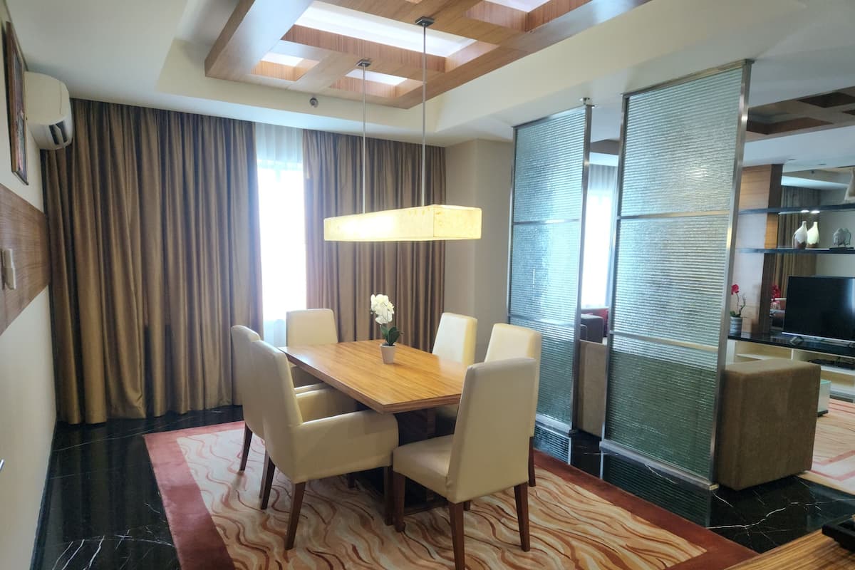 A hotel room with a wooden dining table, four chairs, a potted plant, and a large window with curtains.