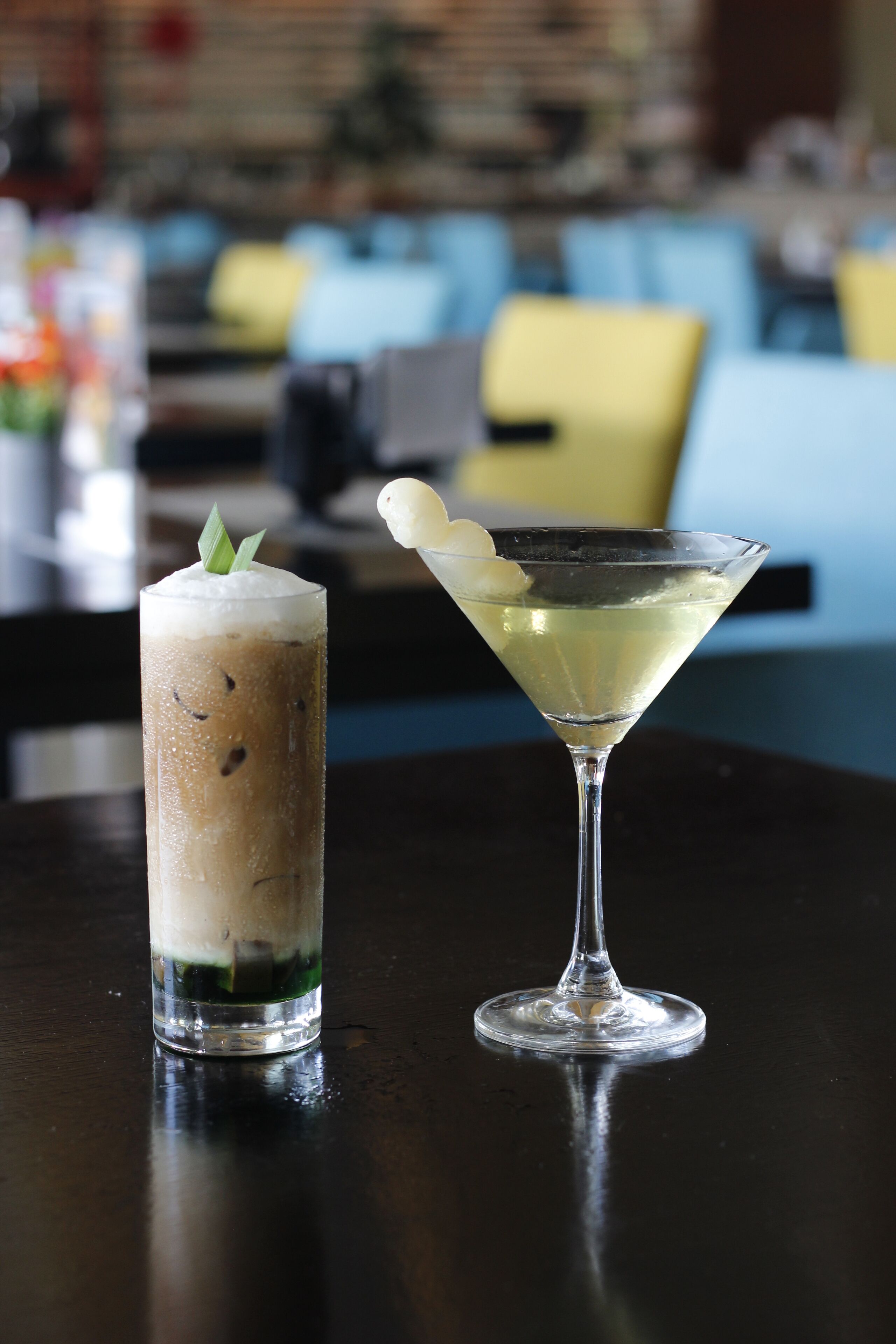 Twee drankjes op een tafel, één in een hoog glas en één in een martini-glas, met een vage achtergrond van een bar.