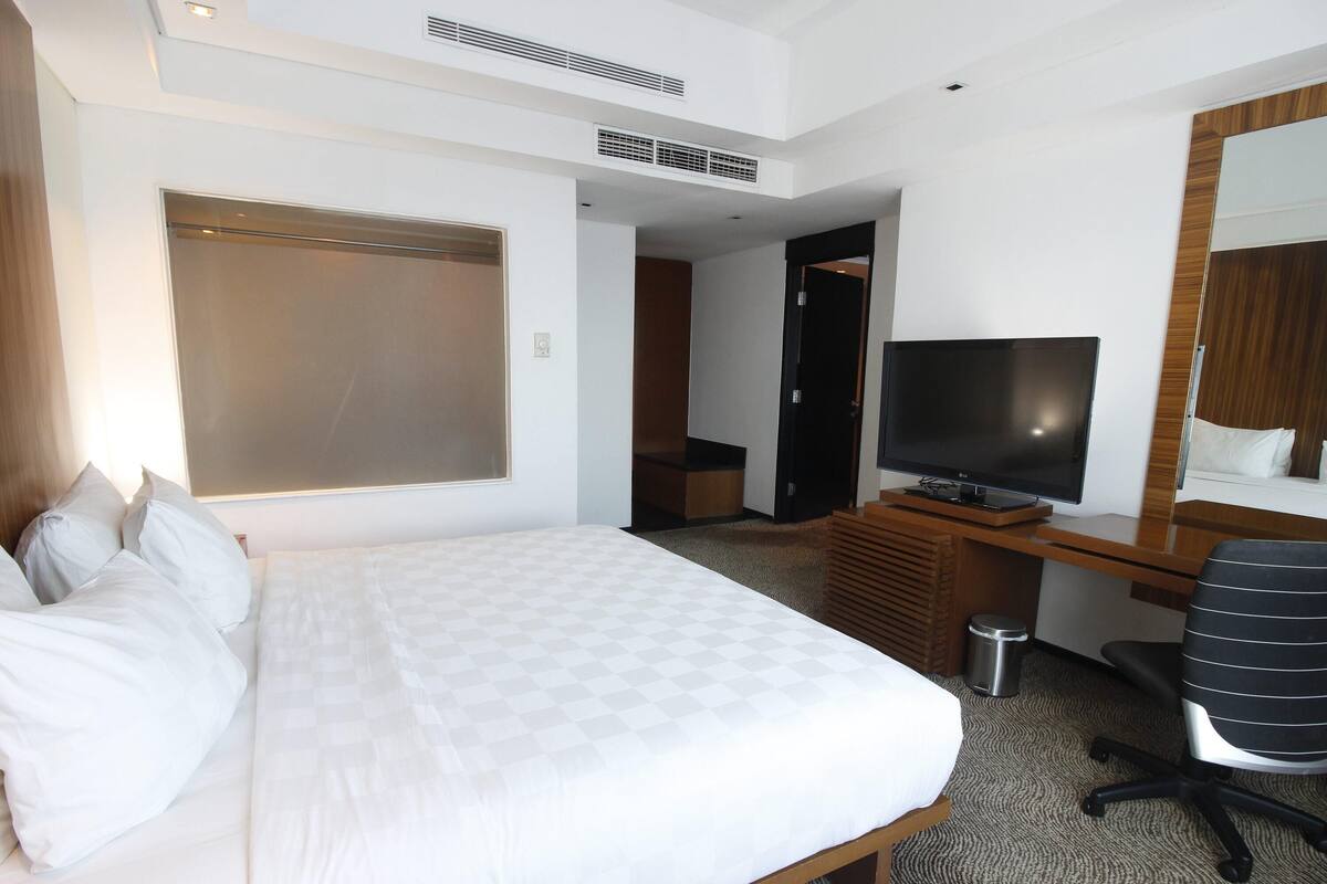 A hotel room with a large bed, a desk with a television, a mirror, and a door leading to another room.