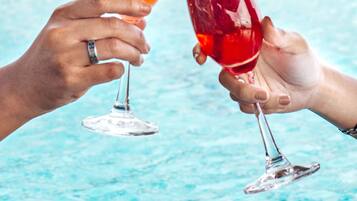 Two hands holding cocktail glasses with straws, one with a red drink and the other with a pink drink, by a pool with a building in the background.