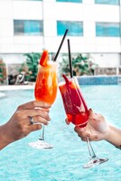 Dos manos sosteniendo copas de cóctel con pajitas, una con una bebida roja y la otra con una bebida rosada, junto a una piscina con un edificio al fondo.