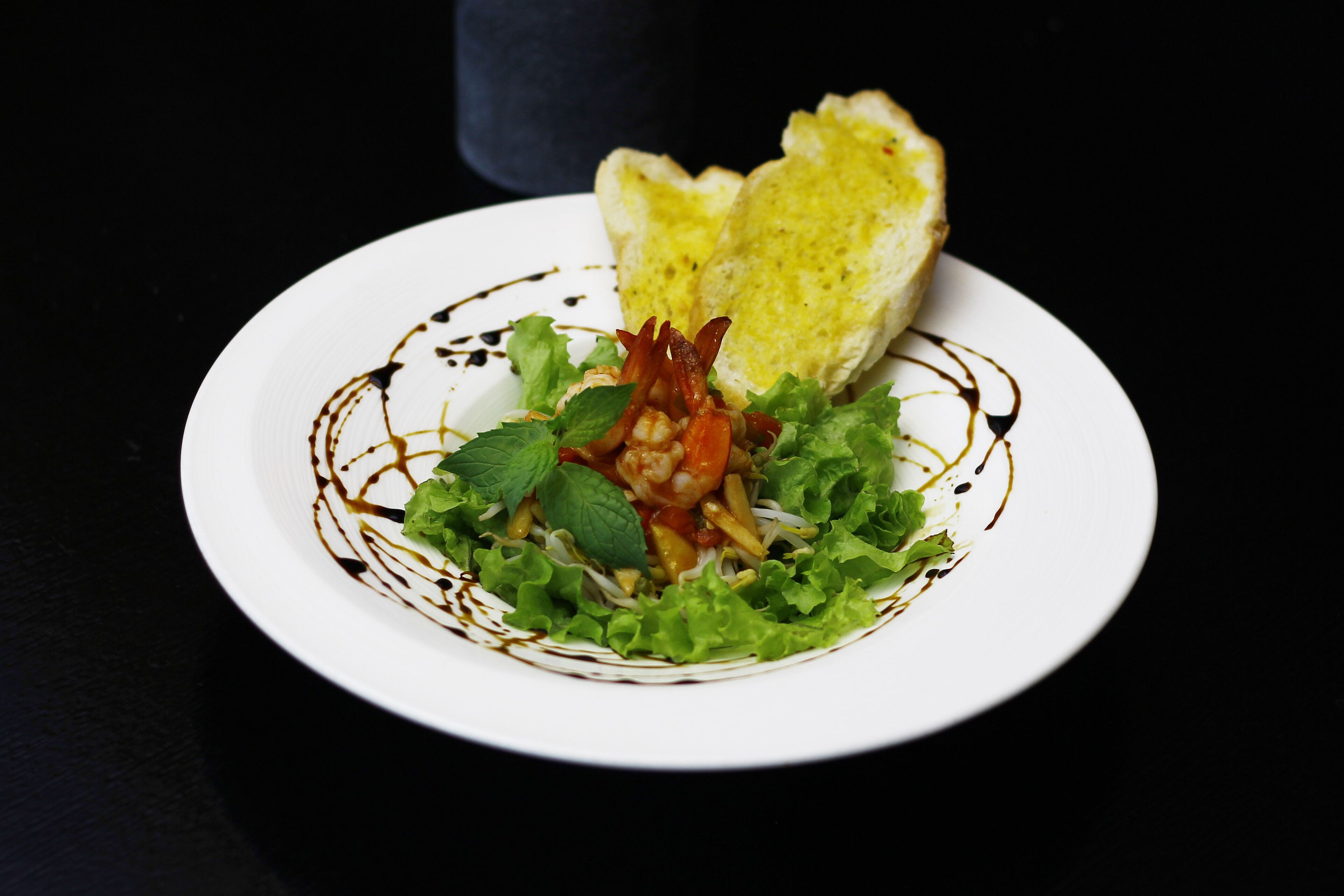 Une assiette blanche contenant une salade, un morceau de pain et une sauce foncée, le tout sur un fond noir.