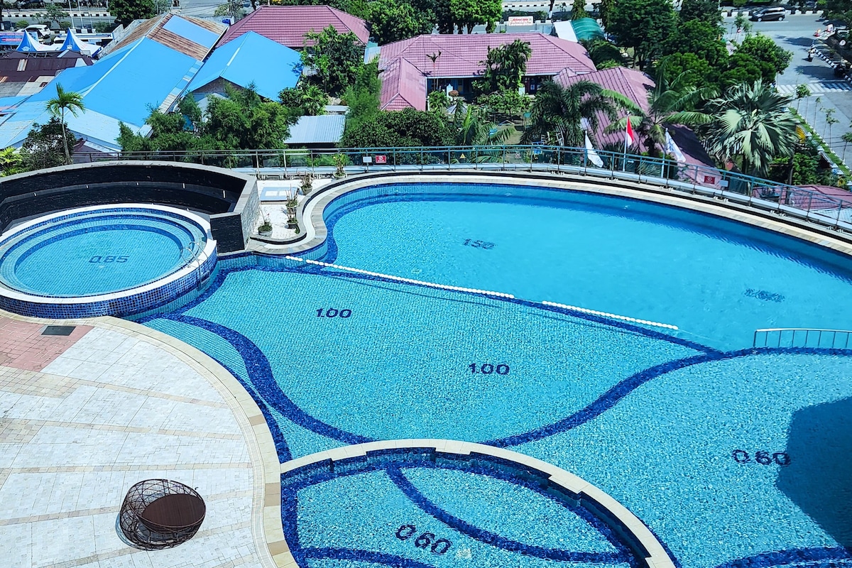 A large, multi-level swimming pool with clear water, surrounded by a tiled deck and a palm tree.