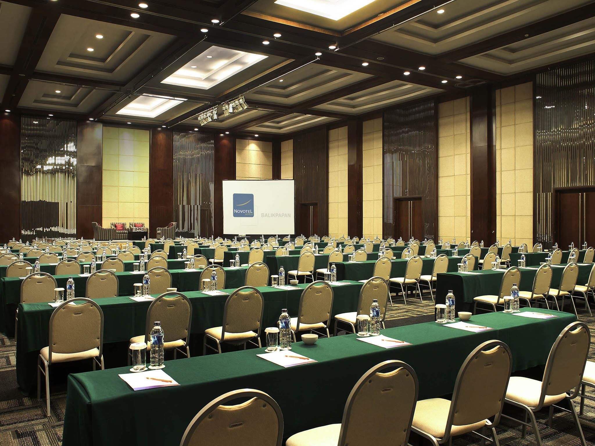 A conference room with rows of tables and chairs, a projection screen, and a large screen displaying the Novotel Balikpapan logo.