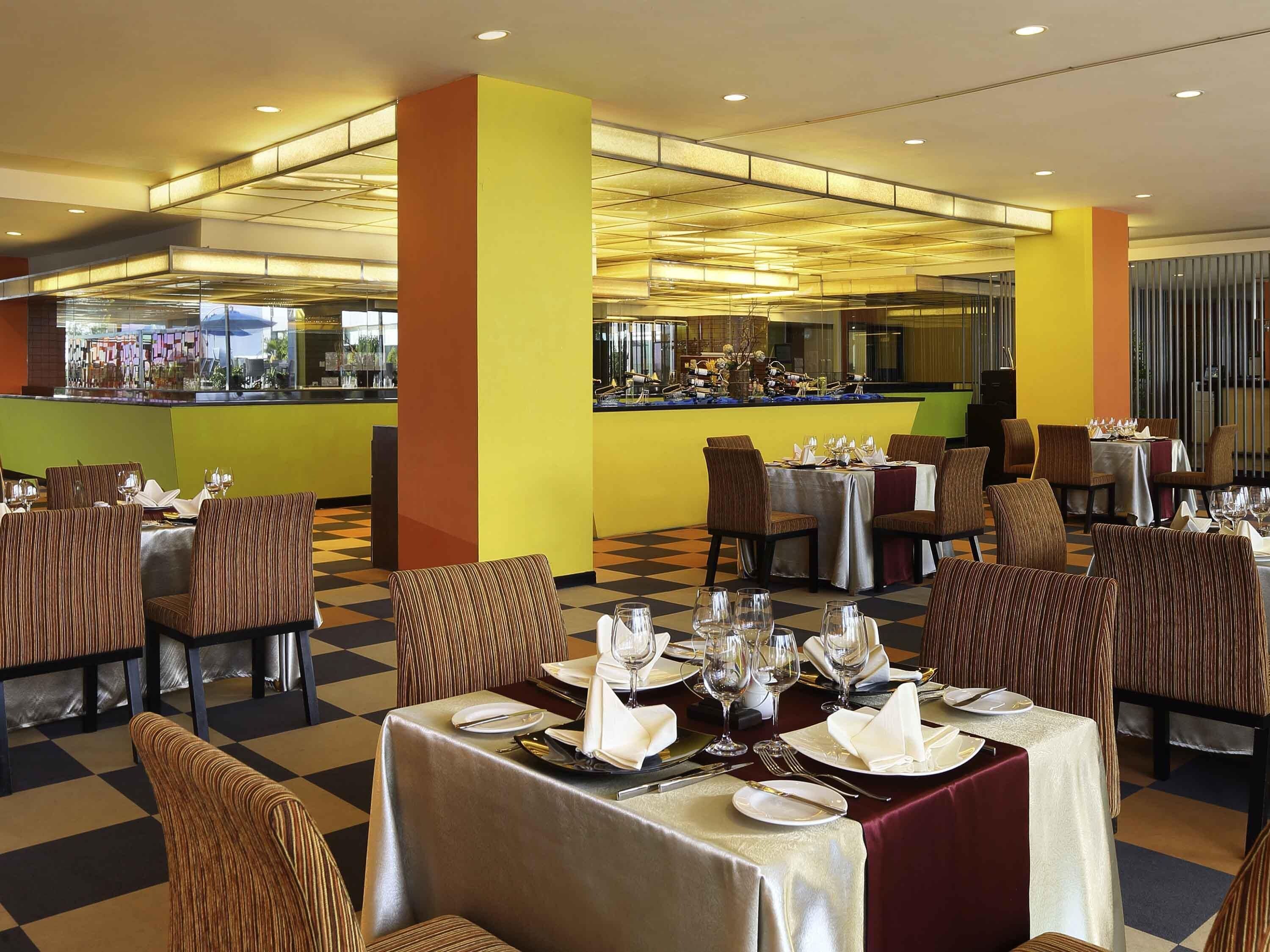 Een restaurant met meerdere tafels klaar voor het dineren, een bar en uitzicht op de keuken.