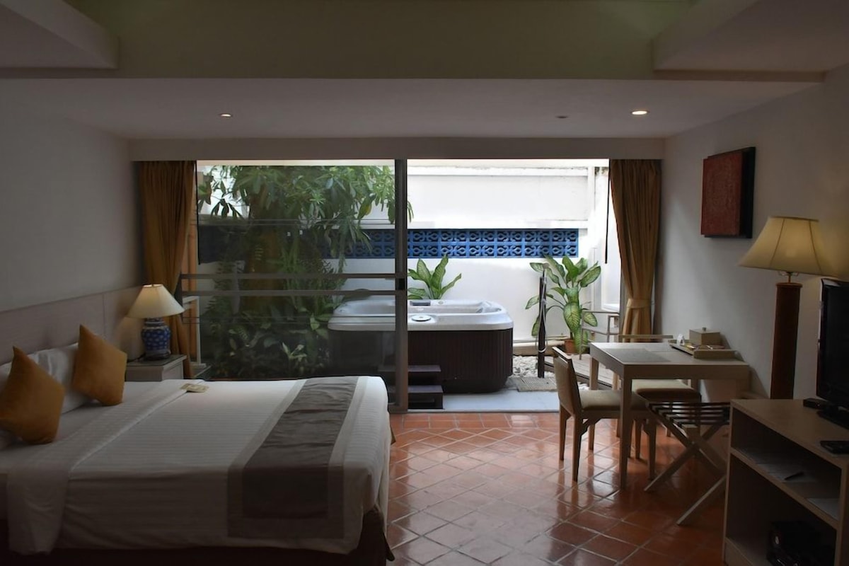 Habitación de hotel con cama, escritorio, silla, televisor, bañera de hidromasaje y balcón con plantas.
