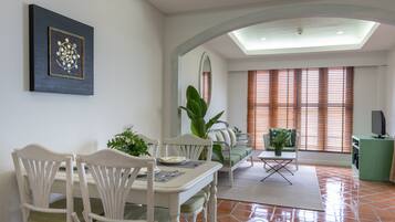 Un coin repas comprenant une table, des chaises et un canapé. Une grande fenêtre avec des stores et une plante d’ornement se trouvent dans un coin de la pièce.