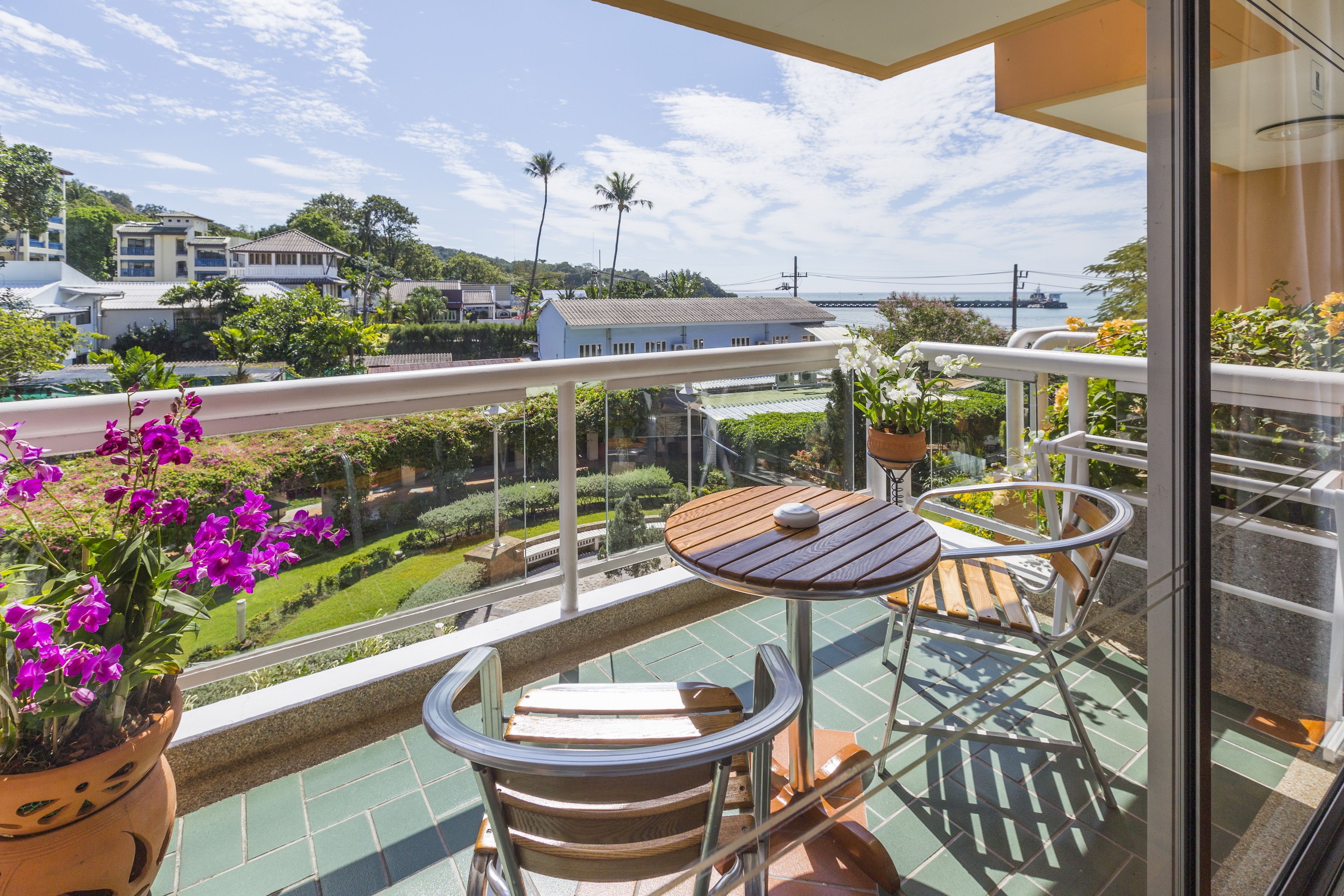 A balcony with a wooden table and chairs, a potted plant with purple flowers, and a view of a coastal area with buildings and greenery.