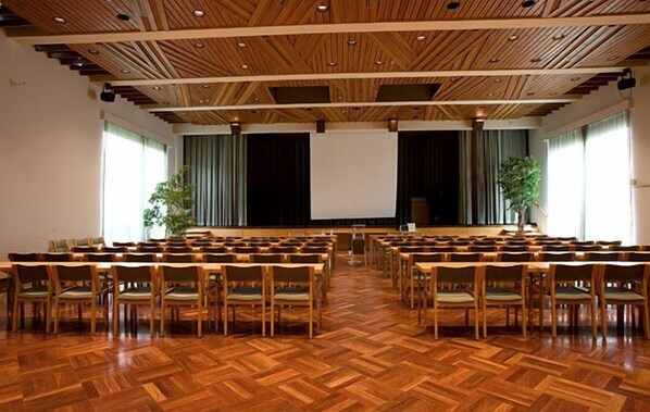 Ein großer Saal mit Holzboden und Holzbalkendecke, mit in Reihen angeordneten Stühlen vor einer Bühne.