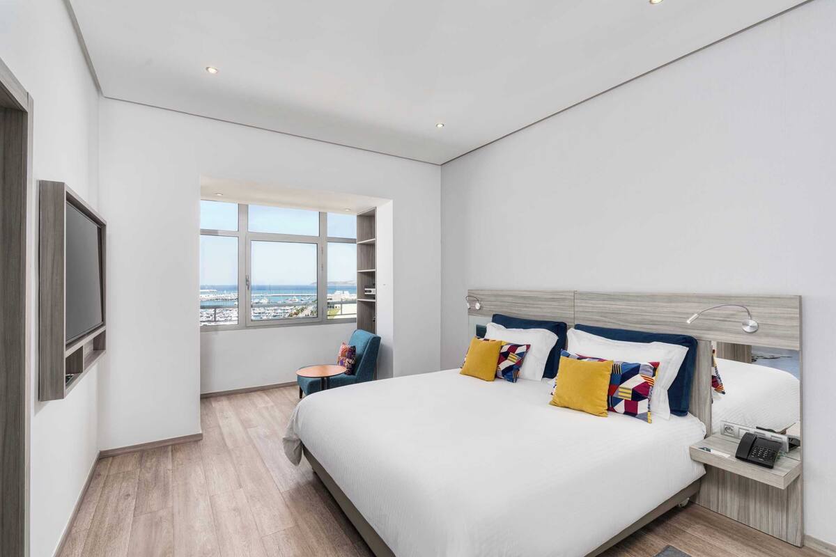 Una habitación de hotel moderna con una cama grande, un televisor, una mesita y un ventanal con vistas a la ciudad.