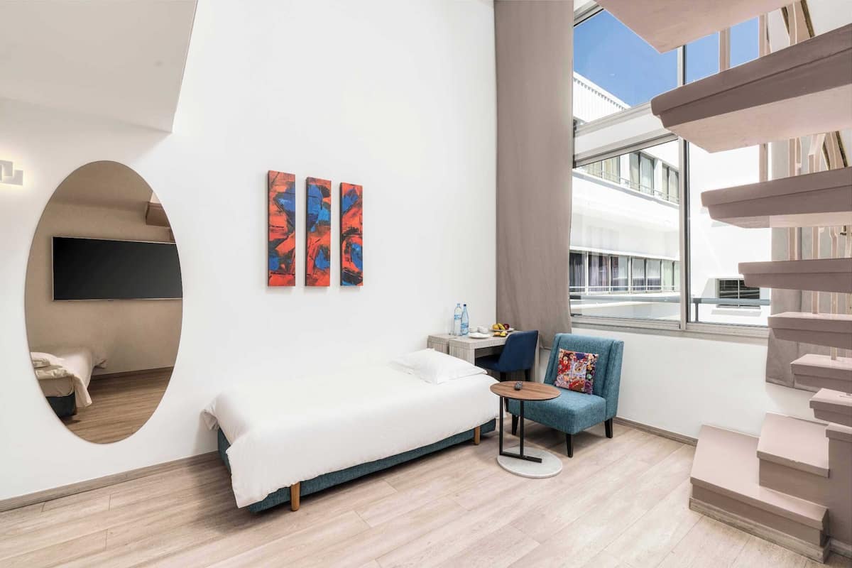 Una habitación de hotel moderna con una cama, una mesita, una silla y un ventanal con vistas del edificio.