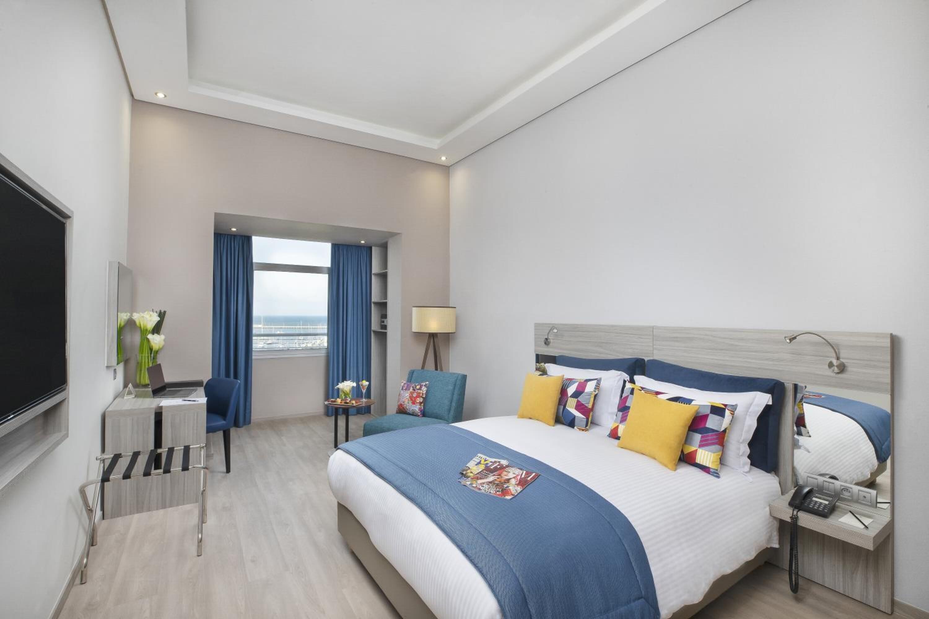 Une chambre d’hôtel avec un grand lit, un bureau avec un ordinateur, une chaise, une petite table avec une lampe et une vue sur la mer.