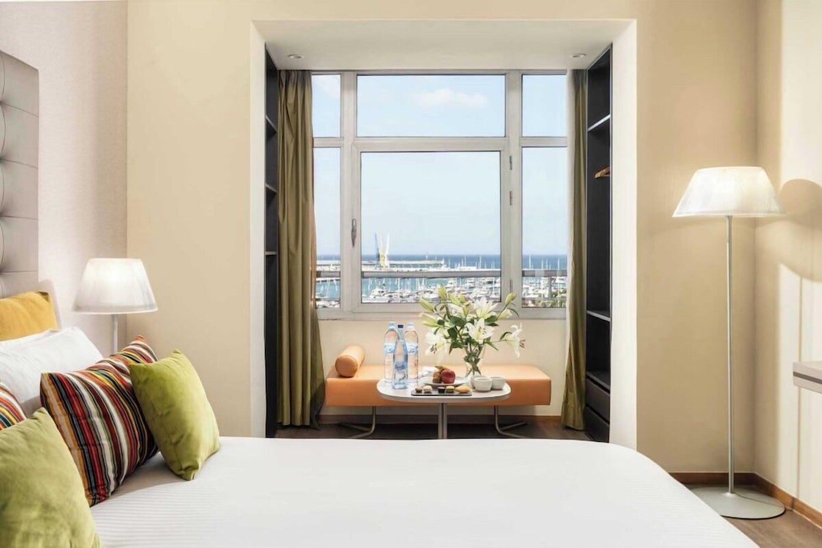 A hotel room with a bed, a bedside table with a vase and water bottles, a window with a view of the marina, and a floor lamp.