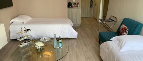 Habitación de hotel con dos camas, una mesa de vidrio con aperitivos y bebidas, una silla y una pequeña cocina en el fondo.