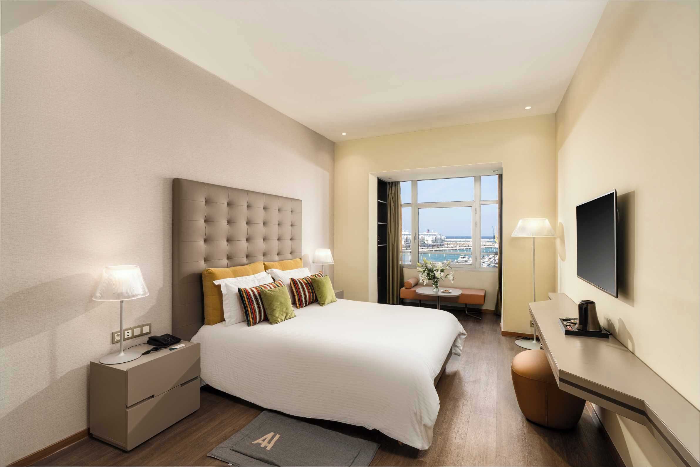 A modern hotel room with a large bed, bedside lamps, a desk, a flat-screen TV, and a view of the city through a window.