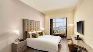 A modern hotel room with a large bed, bedside lamps, a desk, a flat-screen TV, and a view of the city through a window.