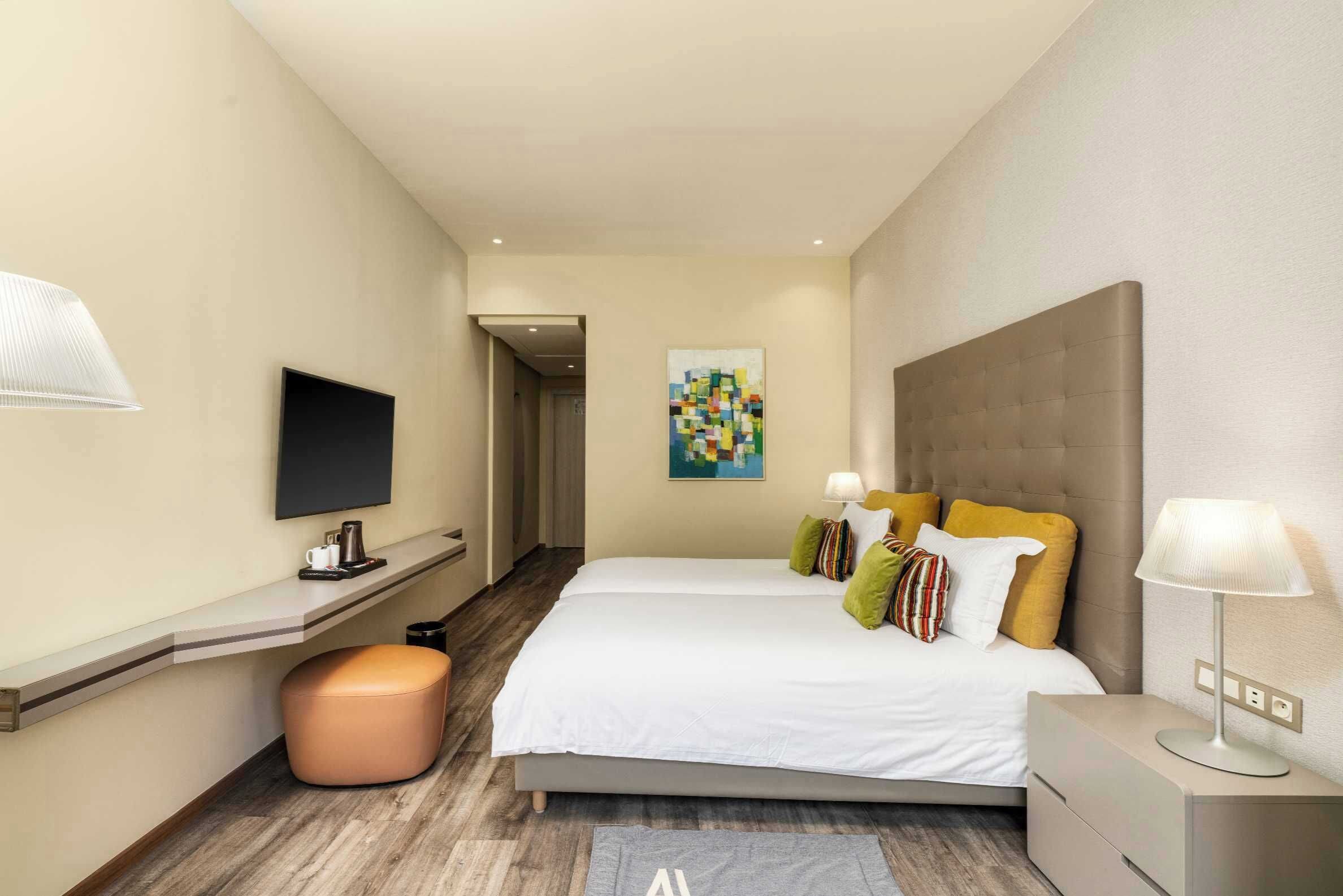 A modern hotel room with a large bed, a flat-screen TV, a desk with a lamp, and a painting on the wall.
