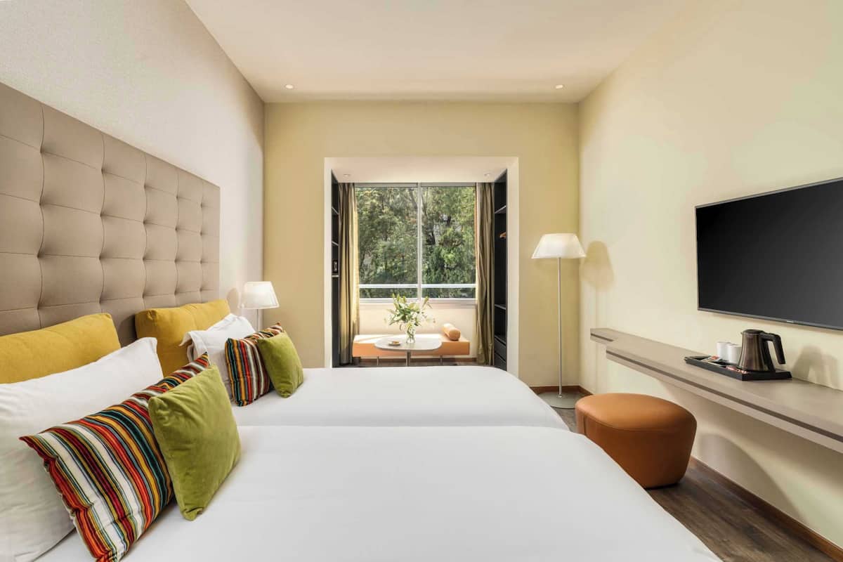 Habitación de hotel con una cama grande, televisión, escritorio con lámpara y ventana con vista a árboles.