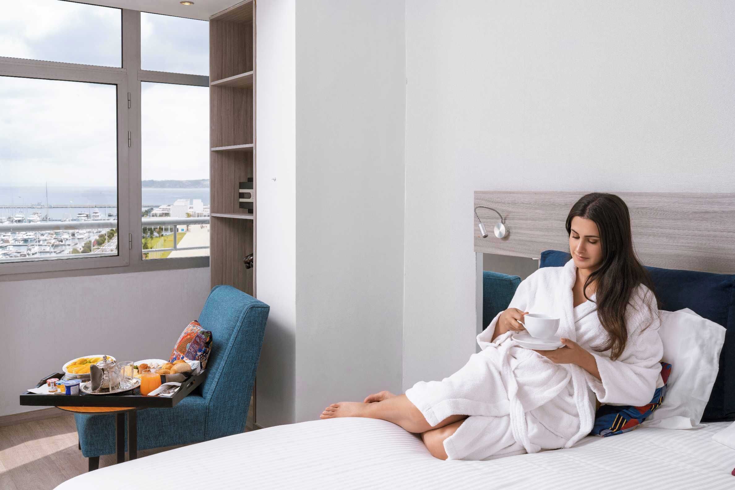 Habitación de hotel con una cama, un sillón azul, una bandeja con alimentos para el desayuno y vistas a la ciudad a través de amplias ventanas.