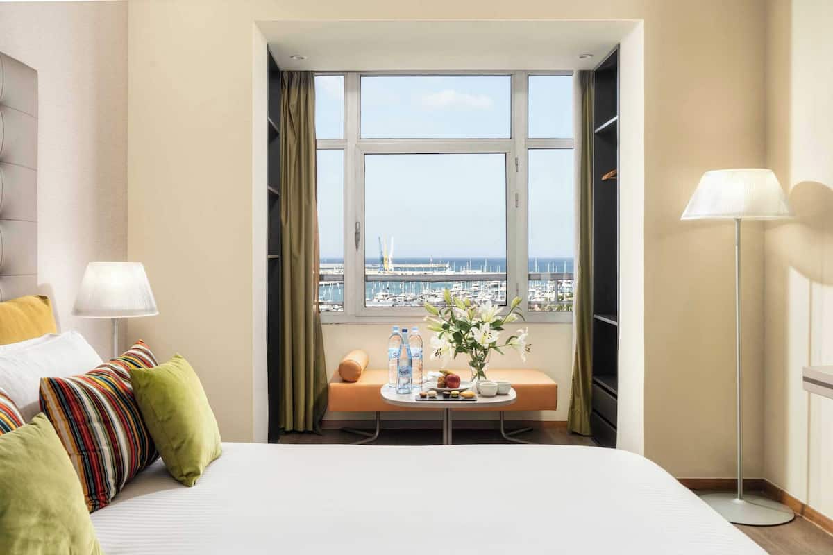 Habitación de hotel con una cama grande, una mesita de noche con un florero, una ventana con vista al puerto deportivo y una lámpara de pie.