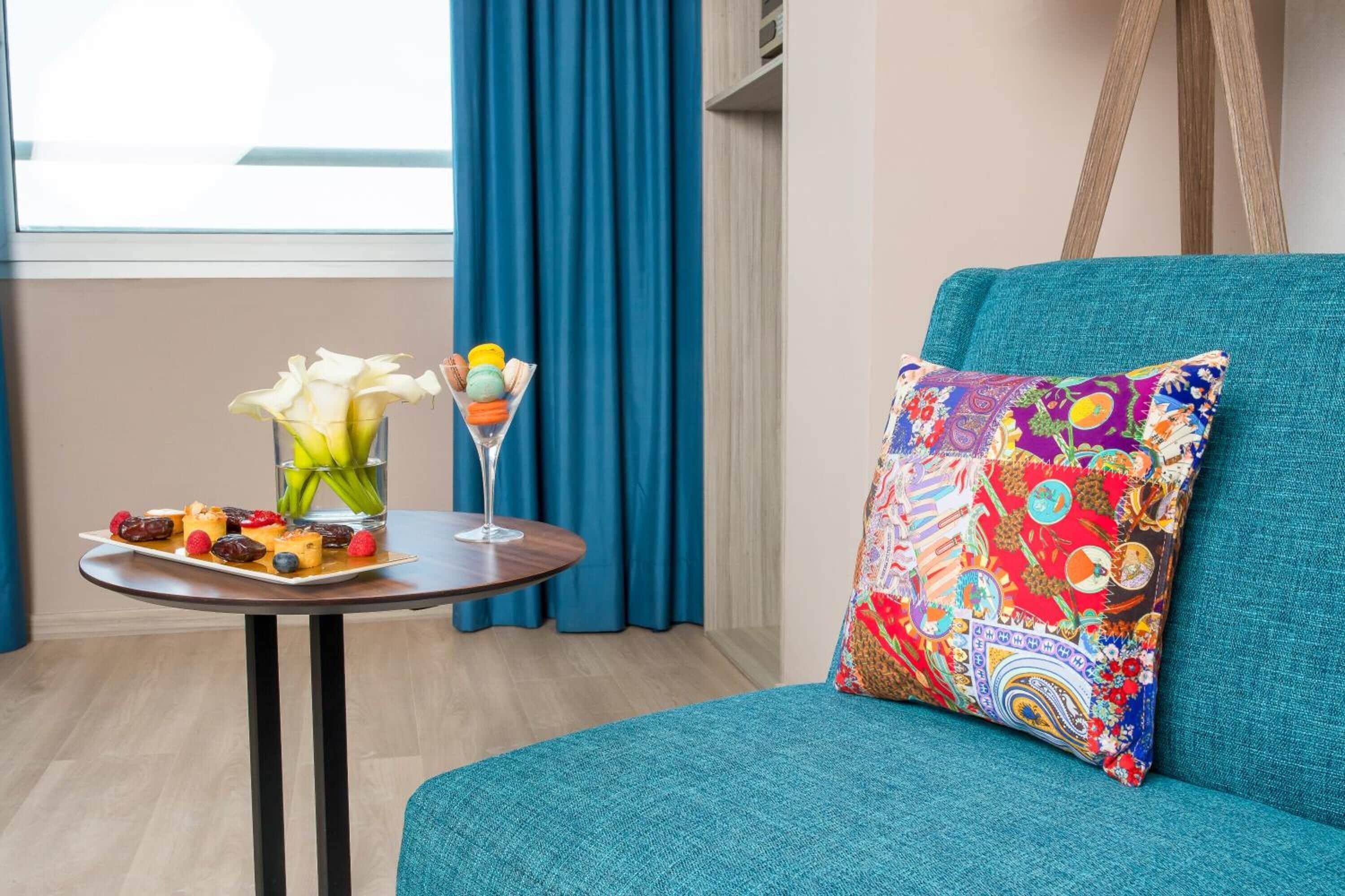 A teal sofa with a colorful pillow, a small round table with a glass of drink and a plate of food, and a vase with flowers.