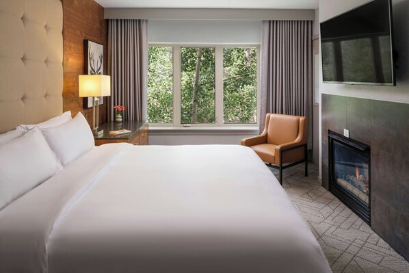 A hotel room with a large bed, a chair, a TV, a fireplace, and a window with curtains.