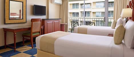 A hotel room with two beds, a desk, a chair, a television, and a balcony with a view of the building.