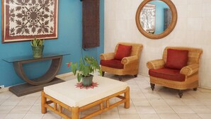 Una habitación con pared azul, una obra de arte floral grande, un espejo redondo, dos sillas de mimbre con cojines rojos, una mesa de madera y una planta en maceta.