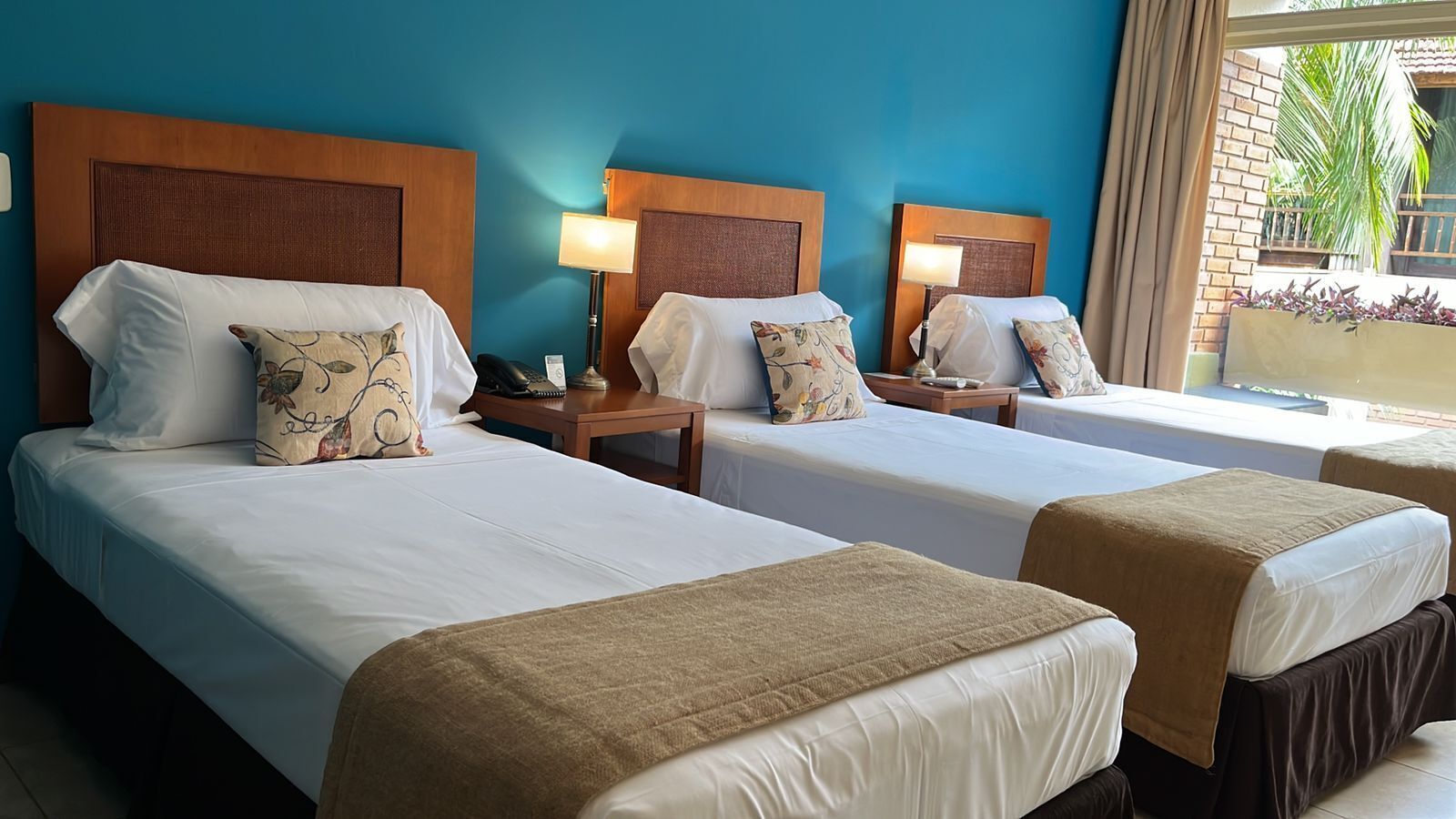 A hotel room with two beds, each with a wooden headboard, white linens, and decorative pillows. There are bedside tables with lamps and a view of the outdoors through a window.