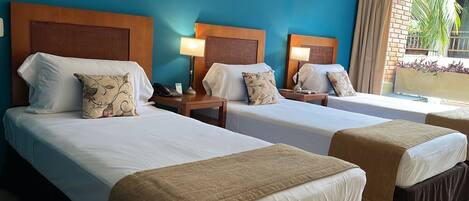 A hotel room with two beds, each with a wooden headboard, white linens, and decorative pillows. There are bedside tables with lamps and a view of the outdoors through a window.