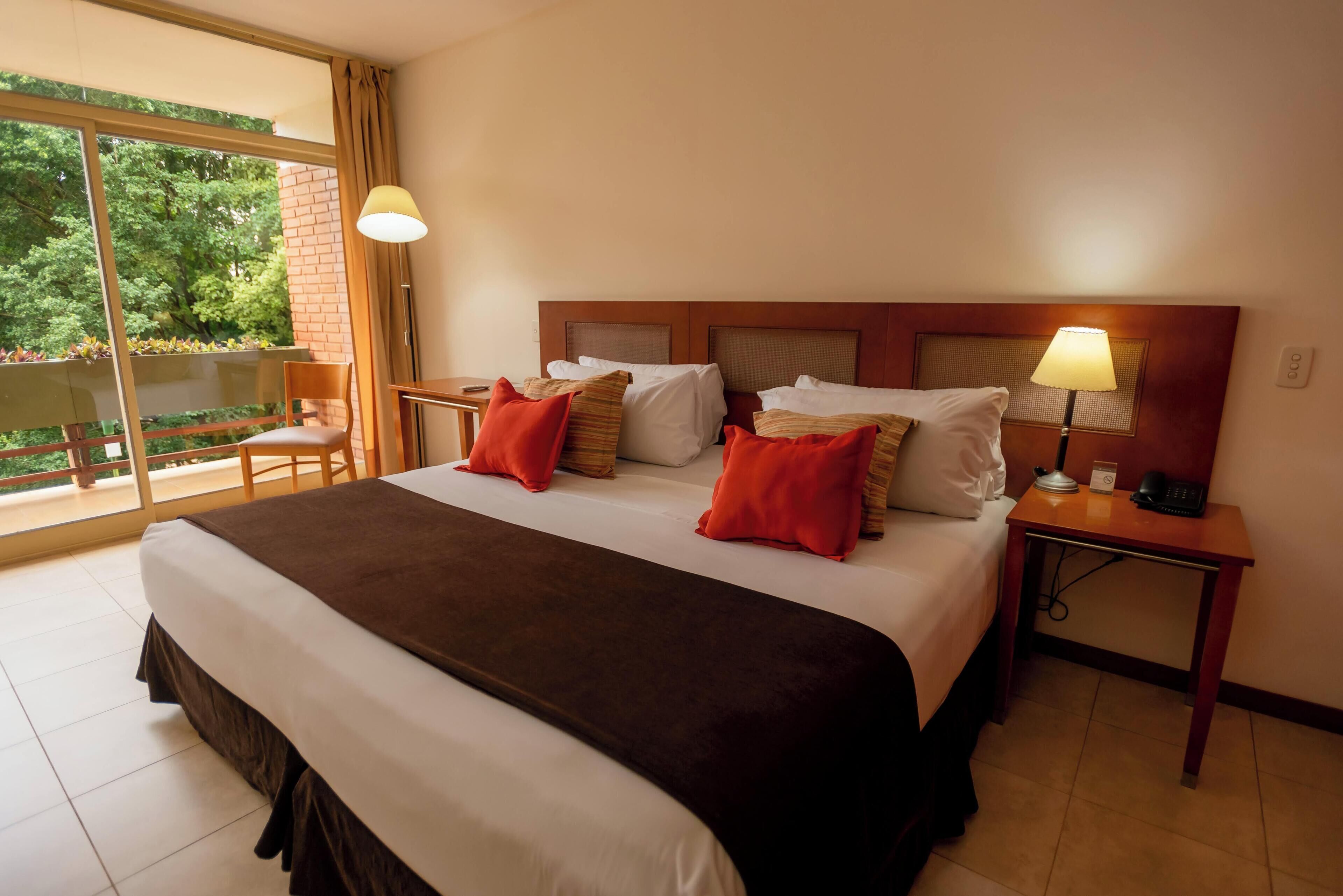Habitación de hotel con una cama grande, dos lámparas de noche, una mesita de noche y vistas a zonas verdes por la ventana.