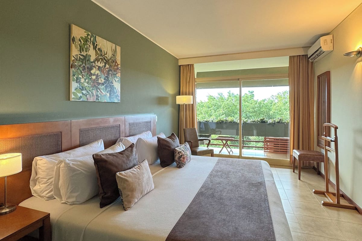 Une chambre d’hôtel avec un grand lit, un bureau, une chaise et une vue sur la verdure environnante.